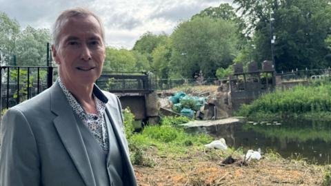 A smiling David King looking straight at the camera. He is wearing a grey suit and is standing on grass in front of brickwork which has collapsed into a river.