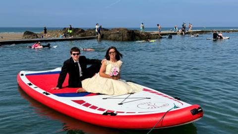 A man in a dark suit and a woman in a cream-coloured dress holding a posy of flowers sit on a paddleboard floating on Bude Sea Pool with swimmers behind them.