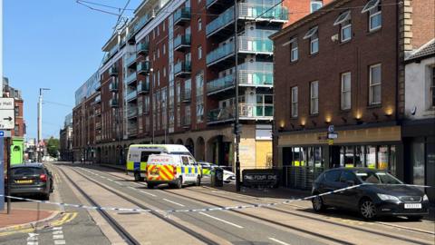 Blue and white police tape stretches across a city centre street. behind the tape, two police vans and a police car are parked , blocking the junction to a side street. The bar is a three-storey red brick building with floor to ceiling windows at ground level and two rows of four standard windows on the floors above. The next building along the street is a modern seven-storey block of apartments with glass-fronted balconies. It is a sunny day with a blue sky.