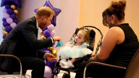 Prince Harry in a dark suit sits besides a little girl wearing a light blue dress. A woman in a black top sits next to the pair. 