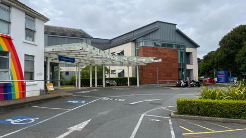 Guernsey's Princess Elizabeth Hospital - A white building with a ranbow painted on it. Next to it is a large criss crossed awning, above a sign which says Le Vauqueidor Entrance. 