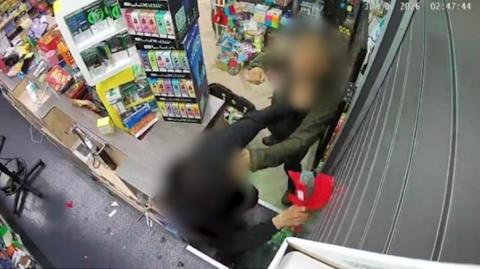 A CCTV image shows a shop assistant being assaulted inside a convenience store in Liverpool. A man whose face is blurred can be seen throwing a punch over the counter at the person working behind it.