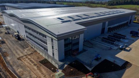 An aerial view of a large grey warehouse taken from the corner of the building. Strips of windows can be seen on the building and some solar panels on the roof. The sun is reflecting back off the roof. To the front of the building exposed soil can be seen, showing the site is not yet complete.