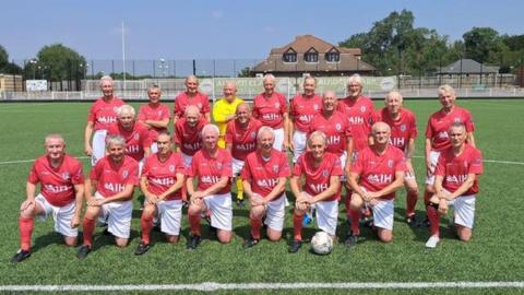 The England over-75s football team are seen on the pitch in a squad formation wearing red tops and white shorts with red football socks