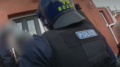Body-cam footage shows a police officer from behind, dressed in protective gear, stood outside a front door
