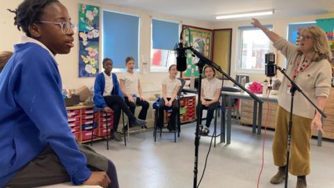 A group of children singing in a modern day classroom with their music teacher as part of the project.