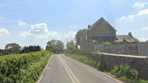 A country A road goes around a bend with a stone house on the right and the hedge bordering a field on the left. It is a sunny day with a few clouds in the sky.