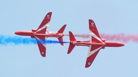 Two Red Arrows fighter planes, with red livery and a white triangle and red, white and blue bullseye symbol on their undercarriages, cross paths in the air while billowing out blue and red smoke 