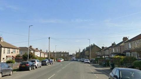 A wide suburban road, populated with parked cars and houses on both sides of the road. It is a bright and sunny day.