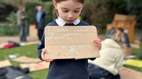 A little girl in her school uniform holds a home-made sign that reads: "I don't want my teachers to leave."