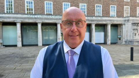 A man wearing glasses and a blue waistcoat and purple tie. 