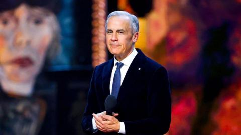 Prime Minister Mark Carney pictured on the stage of the Juno awards in Canada. He is smiling and holding a microphone in his hand. Behind him is a blurred image of Canadian singer Joni Mitchell. He is wearing a suit and a dark blue tie. 