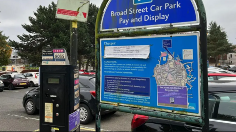 A large but old parking pay and display sign for Broad Street car park can be seen on the right- hand side of the image detailing prices, a map and parking instructions. The metal is rusting. There is a black payment machine next to it and further parking details on its side.