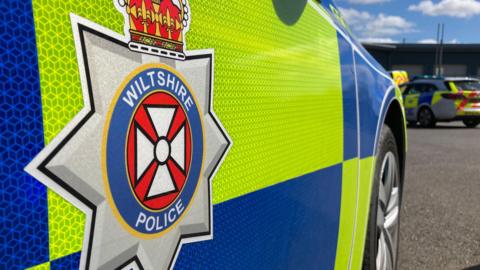 The side of a Wiltshire police car. It is parked in a paved car park.