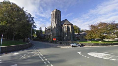 A flat roundabout to the right sits between three roads. Saddle Road runs from right to left, with a large stone church to the righthand side of the walled roads, with trees on the left. It is a sunny day with some whispy clouds.