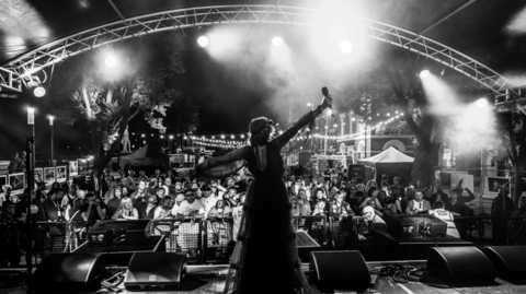 Black and white shot of Hull artist Jodie Langford on stage with crowds of people in front. The performer is holding a microphone in an outstretched arm under an array of stage lighting.