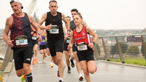 Judith Storm running during the race. She is wearing a red top and black shorts. Running across a bridge.