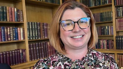 Jenny Lane, who has light brown bobbed hair and is wearing glasses, smiles at the camera. She is wearing a floral blouse and is sitting in front of a wall of bookshelves.