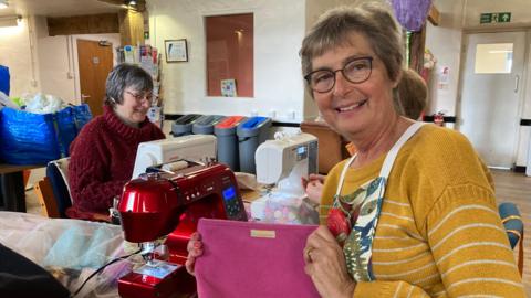 A indoor workspace that appears to be a sewing or crafting room.
A woman wearing a mustard-yellow, long‑sleeved, striped sweater and an apron is holding up a pink fabric item, possibly a handmade pouch or bag.
The fabric piece has a small rectangular label attached near the top.
Another woman is seated at a table, operating a white sewing machine.
Next to that machine, there is a red sewing machine, and various pieces of fabric and supplies are scattered across the table.