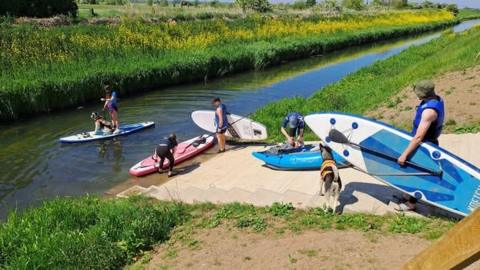 Five paddleboarders at a waterway surrounded by marshland. One person is in the canal on a surfboard with a dog sitting in front of them. Two others are carrying their boards under their arms while walking down a sand-coloured ramp. Another person is on the bank trying to get on to the board. The other is bent over a blue kayak, which is midway down the ramp.