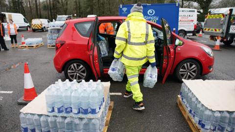 A worker in a hi-vis outfit loads water into a car, with pallets of water either side