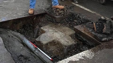 The picture shows a street excavation site. A large open hole in the road surface exposes pipes and a section of granite beneath the tarmac. In the hole is a large stone equal-sided cross.