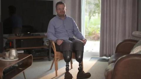 Neil Hopper is sitting in his lounge. He has prosthetic legs and is wearing a blue shirt and a pair of glasses.