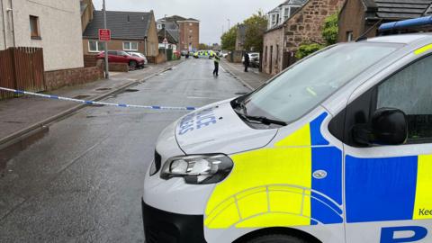 A partial side view of a police van parked in front of a street in Merkinch. The street is lined by different types of properties, including traditional stone-built houses and more modern blocks of flats. The street is wet following a rain shower, and there is police tape stretched across the street next to the van. A police office and another police van are further up the street.