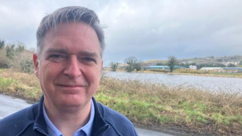 Mr Clelland has short grey hair and is smiling at the camera as he wears a light blue shirt and a dark blue fleece.