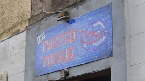 The sign above the door of The Twisted Tongue bar. The name of the bar, written in a style resembling neon lights, is accompanied with a picture of a tongue, tied in a knot protruding from a mouth.