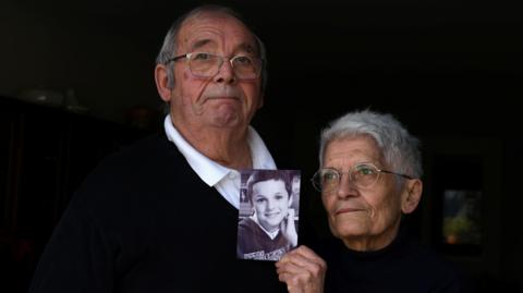 An elderly man and a woman hold up a black-and-white photo of a child