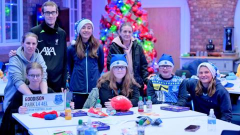 Two Everton players with parents and a grandparent and children, in blue Santa hats, at a table, smiling at the camera 