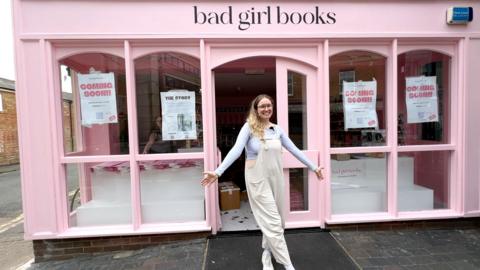A woman stands in front of a pink shop with Bad Girl Books written above the door.