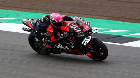 Asprilia motorbike covered in sponsor names. It has some red trim. The rider has a pink helmet and red and black leathers. The bike is leaning to the right as the bike takes a right-hand bend on grey tarmac, with a green and white strip to the biker's left.