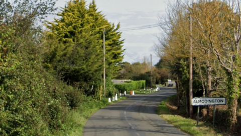 Almodington Lane in West Sussex near Chichester. Pictured is a narrow lane with green trees on either side and a white sign which reads Almodington