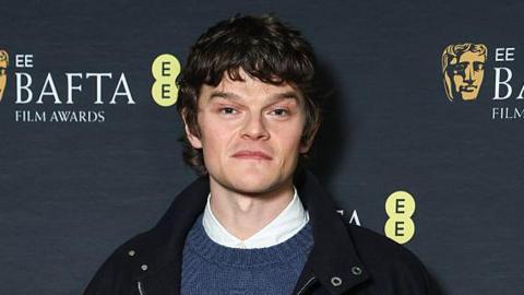 A head and shoulders shot of Robert Aramayo looking at the camera. He has brown hair and is wearing a black jacket, a blue knitted sweater and a white collared shirt. 