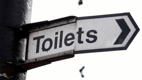 A black and white street sign points to the right, indicating the nearest toilets. The sign is a little bent in the middle, and it's attached to a post.