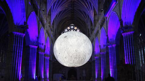 A large spherical sculpture of the moon hangs in the arches of a gothic cathedral at night, with the columns illuminated by purple light.