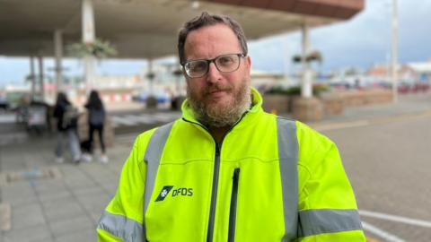 A man wearing a high vis jacket with DFDS logo on the right chest area, background of the harbour is blurred.