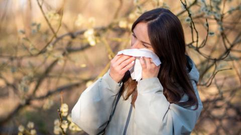 A young woman with long brown hair wearing a grey jacket dabs her eye with tissue, with flowering trees in the background behind her