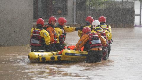 A group of rescue workers, mainly wearing red helmets, push a rescue boat through flood waters with heavy rain falling.