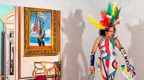 Quilla Constance, dressed in a colourful outfit, with large feathers in her hair. She is holding a paint brush in one hand and a painting pallet in the other. She has on one blue wrist sleeve and one black and white wrist sleeve. She is in a room with a chair and a painting in a frame.