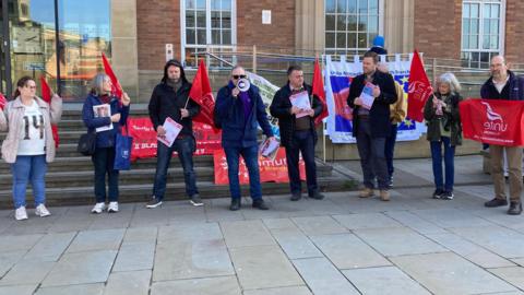 Men and women from the Unite union have flags and banners outside the council house in Derby as they protest against what they claim are cuts to a cancer service.