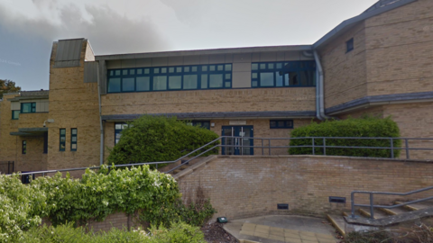 Photo of Shrewsbury Crown Court; it's a modern building, built out of sand-coloured bricks and grey-black-framed windows. In the foreground you can see bushes and shrubs, and steps and hand railings leading up to the court entrance. It's a sunny but overcast day. 