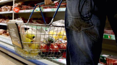 A shopper wearing blue denim jeans holds a metal wire shopping basket with blue handles while looking at packets of meat in a shopping aisle. The basket has bags of fruit and veg in it, including bananas, tomatoes, cucumbers and peppers.