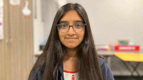 Eyva is smiling and looking at the camera. She is wearing a navy hoody and red and white T-shirt. In the background there are tables and cupboards inside a classroom. 