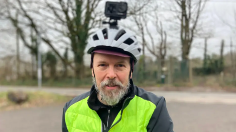 A cyclist wearing a grey helmet with a camera on it is standing in the centre of the frame. He is wearing a reflective jacket. Sparse trees line a road in the background.