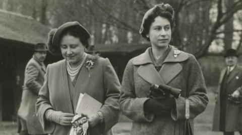 A black and white photograph of Queen Elizabeth, The Queen Mother, and Queen Elizabeth II. Both are wearing long coats and hats. 