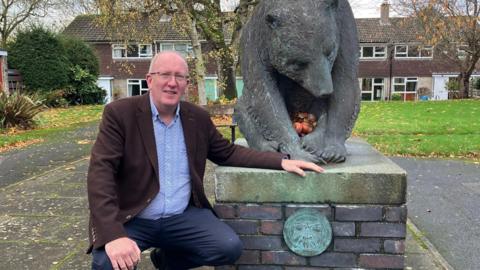 Stephen Fenton kneels beside a large bronze bear sculpture mounted on a brick and stone pedestal in an outdoor setting. The bear is positioned with its head lowered and front paws together, holding several apples. The pedestal features a circular plaque with an engraved design.