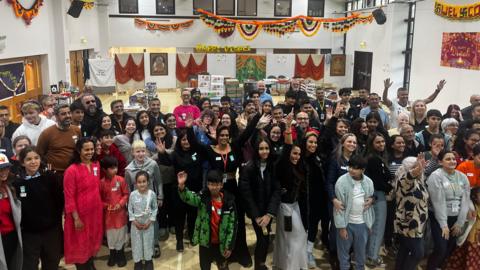 A large crowd of people of all ages raises their arms for a picture in a big hall, decorated with orange, yellow, green and red banners and bunting for Diwali